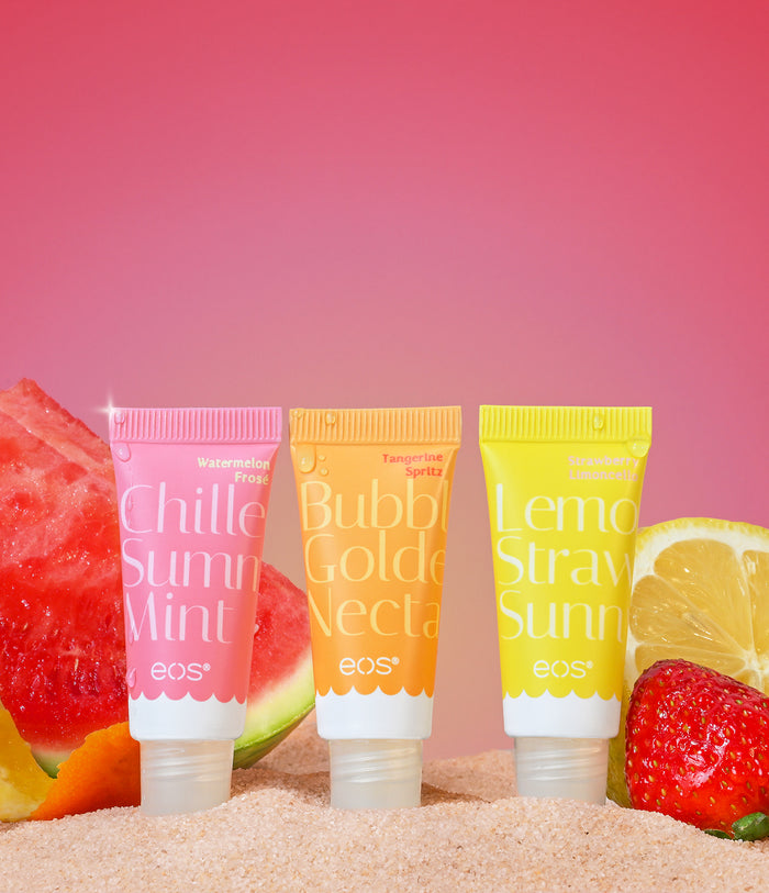 Three eos lip balm tubes with colorful labels on a beach setting with fruits.