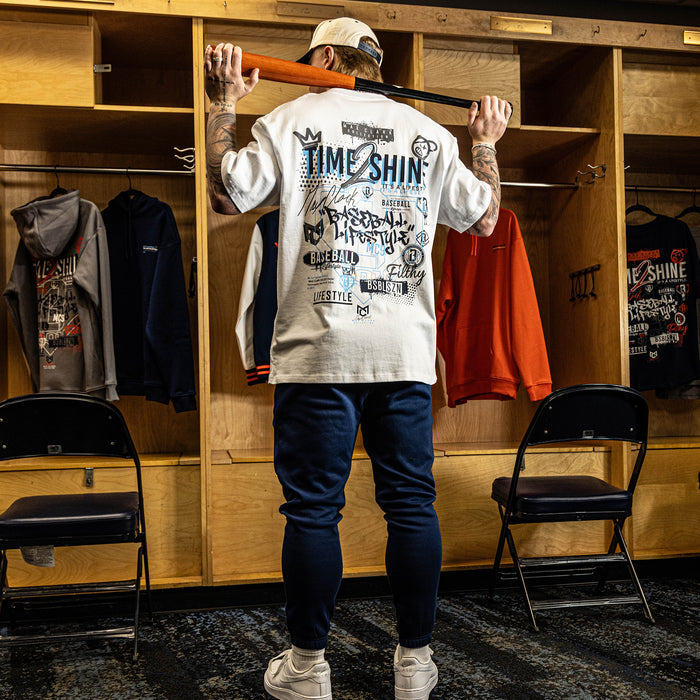 Person in a locker room wearing a white graphic sweatshirt and blue pants, holding a baseball bat.