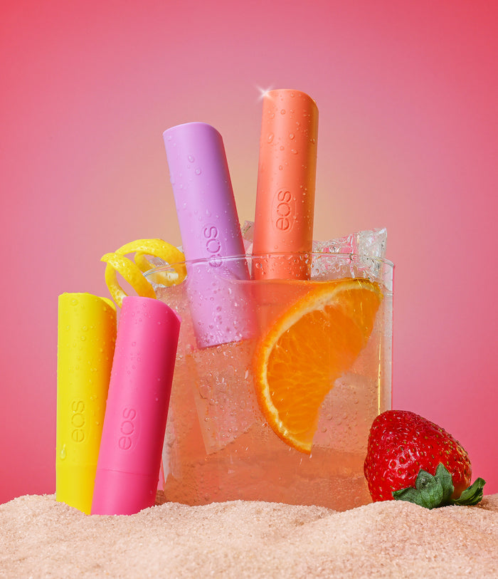 Four EOS lip balms in different colors with a glass of orange juice and a strawberry on a pink background.