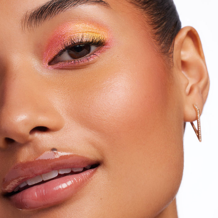Shimmery pink-to-gold chrome eyeshadow blended across the eyelid, showcasing Bliss, the ColourPop Chrome Liquid Shadow, on a close-up model with full lashes, glossy nude lips, and a gold geometric hoop earring in bright studio light.