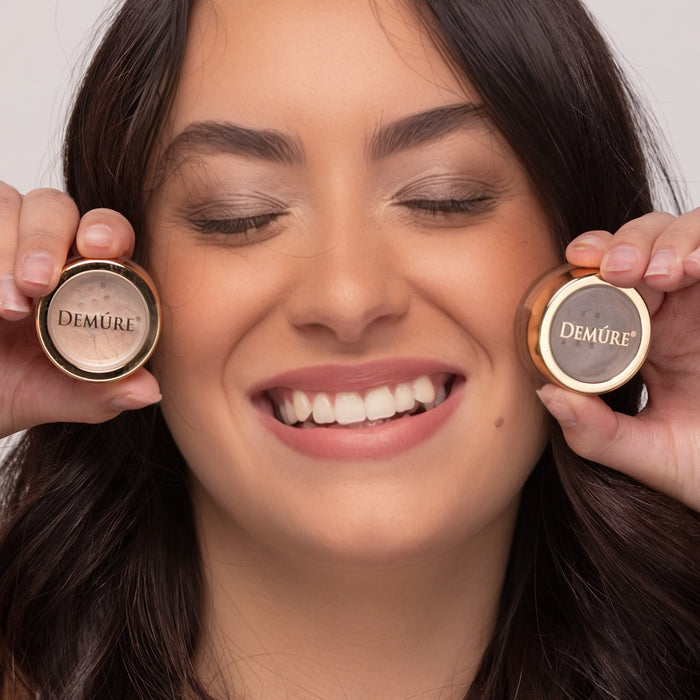 Woman wearing Demure Brown Liner & Golden Sand eye colors, holding the  two 'Demure' makeup jars near her face