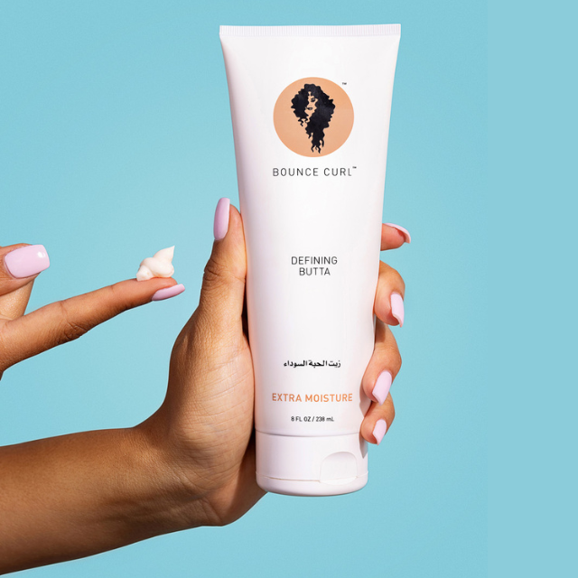 Hand holding a Bounce Curl tube; dollop of product on fingertip, teal background.