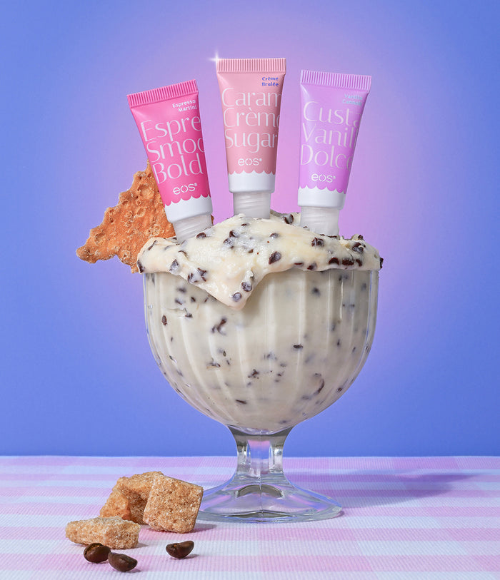 Three tubes of Buxom lip balm in a dessert glass with ice cream and cookies on a pink and purple striped background.