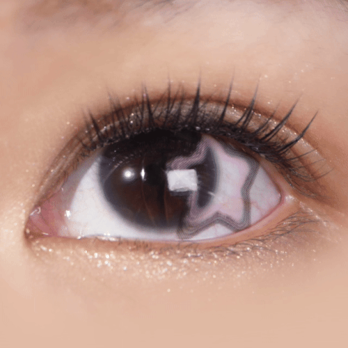 A close-up of a human eye with long lashes wearing EyeCandys Pink Star contacts by EyeCandys, shows a star-shaped reflection in the pupil and subtle shimmer on the eyelid.