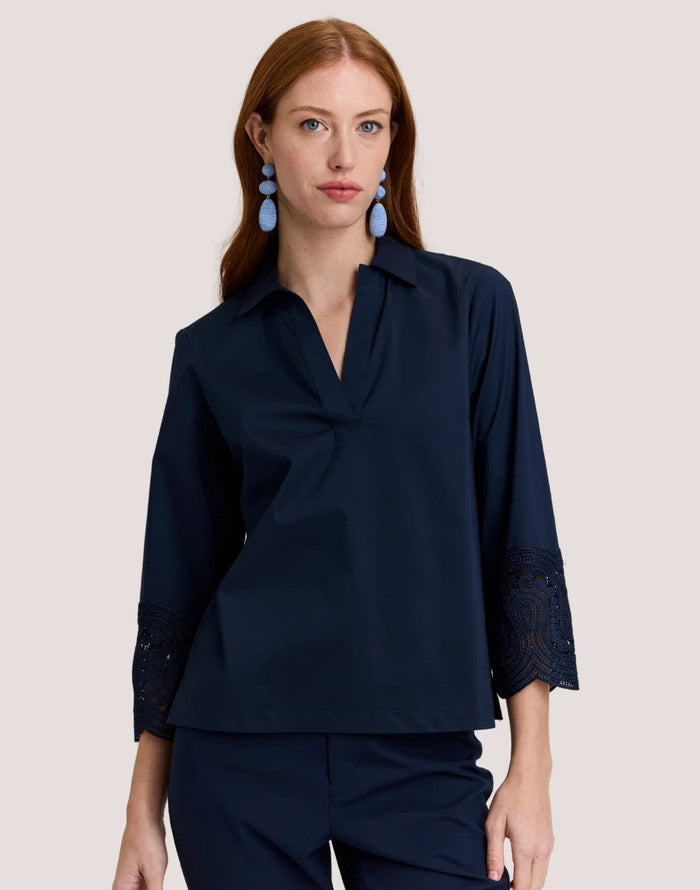 A woman with long red hair wears the HINSON WU MONA Bracelet Sleeve Stretch Polished Cotton Lace Top in navy—a comfortable, stretch cotton blouse featuring three-quarter sleeves with lace accents—paired with navy pants. She accessorizes with blue statement earrings and stands against a plain light background.
