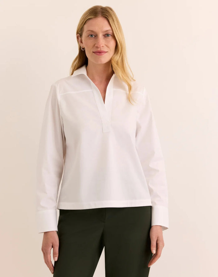 A woman with long blonde hair models the HINSON WU Monica Long Sleeve Button Back Top in white—a collared stretch polished cotton blouse featuring a split neckline and long sleeves—paired with dark green pants, standing against a plain light background.