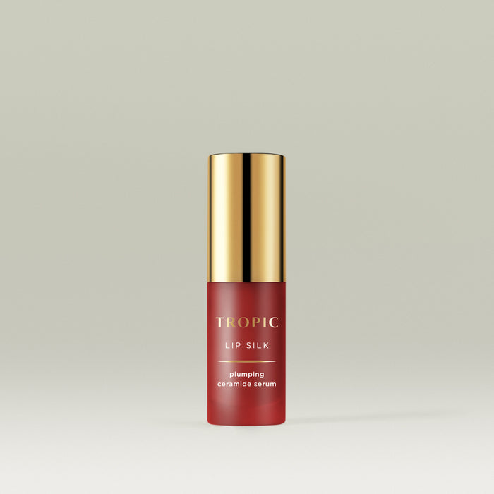 A sleek red bottle of Tropic Lip Silk plumping ceramide serum with a shiny gold cap, standing upright against a soft neutral background.