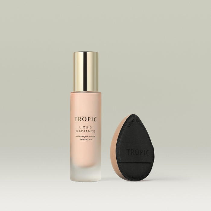 A frosted glass bottle of Tropic Liquid Radiance adaptogen serum foundation in shade 1 with a gold cap, displayed beside a black teardrop-shaped velvet blending cushion with a beige base.