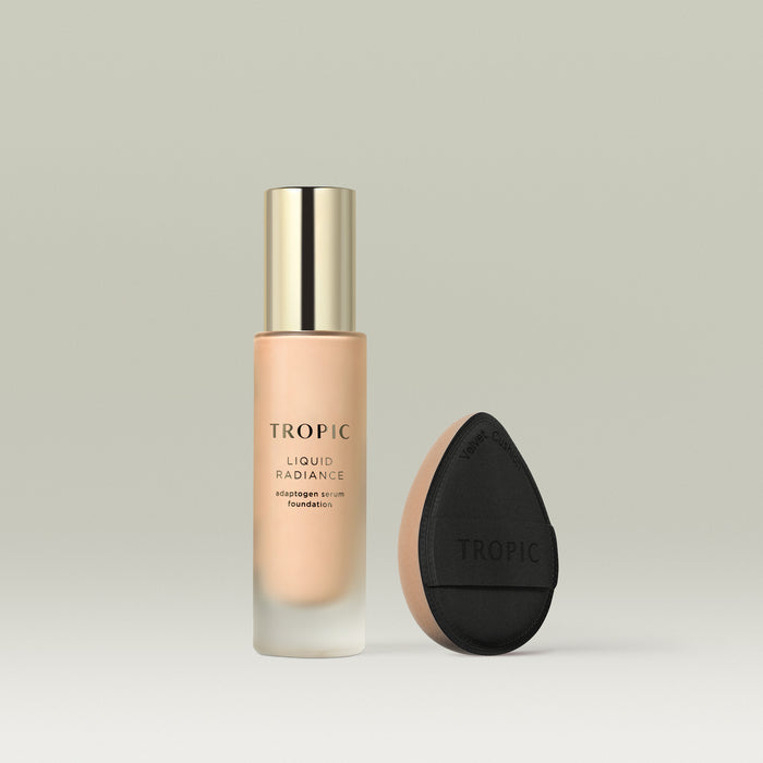 A frosted glass bottle of Tropic Liquid Radiance adaptogen serum foundation in shade 2 with a gold cap, displayed beside a black teardrop-shaped velvet blending cushion with a beige base.