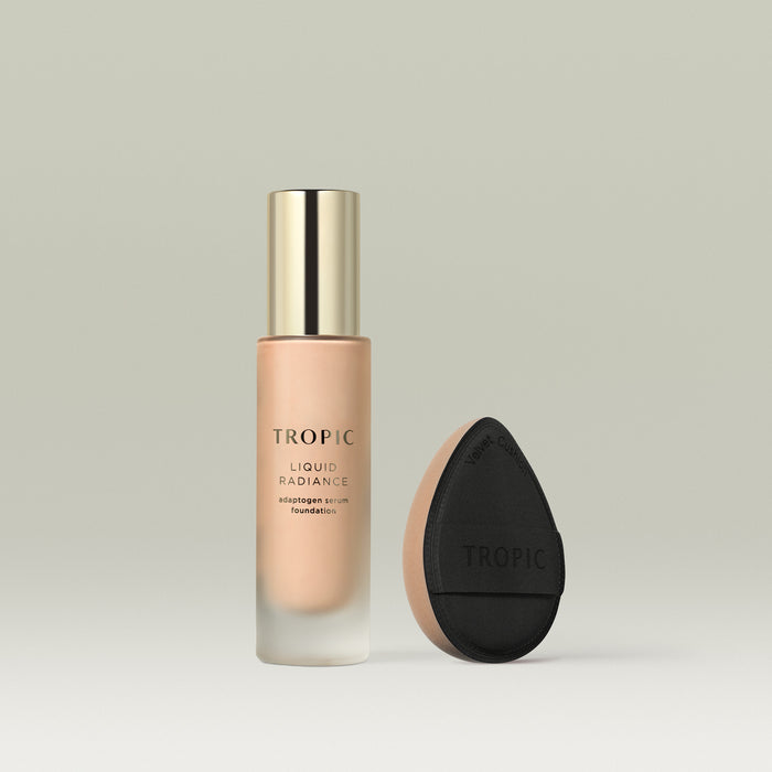 A frosted glass bottle of Tropic Liquid Radiance adaptogen serum foundation in shade 3 with a gold cap, displayed beside a black teardrop-shaped velvet blending cushion with a beige base.