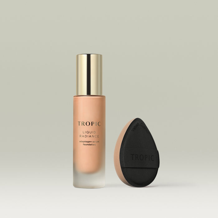 A frosted glass bottle of Tropic Liquid Radiance adaptogen serum foundation in shade 4 with a gold cap, displayed beside a black teardrop-shaped velvet blending cushion with a beige base.