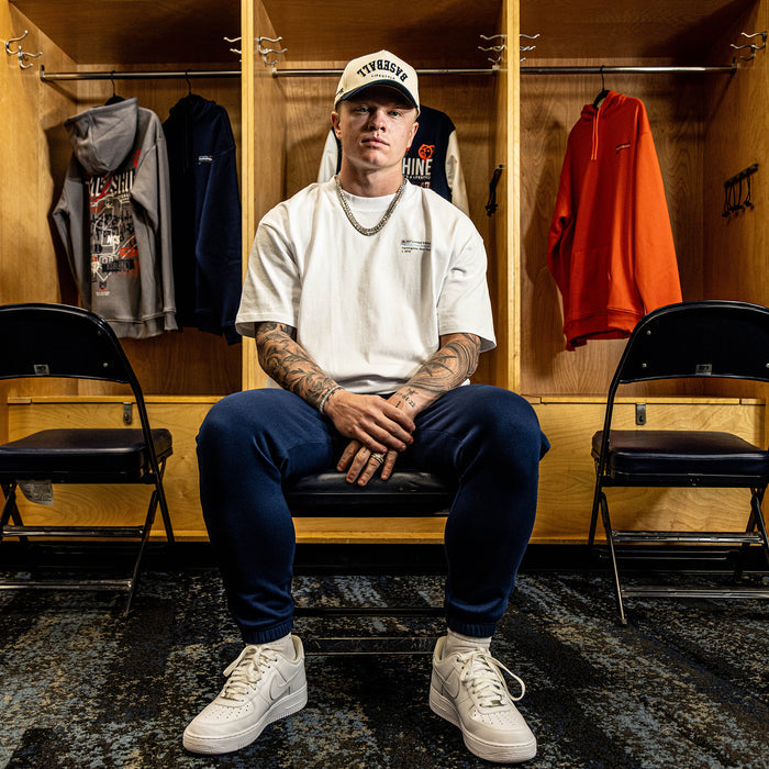 Person sitting in a locker room wearing a white t-shirt, blue pants, and a white cap.