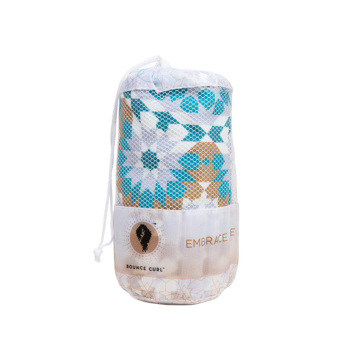 A Bounce Curl micro-fiber towel with a teal, gold, and white pattern rolled up and stored in a white mesh drawstring bag