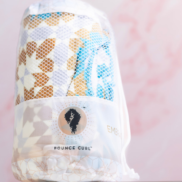 Bounce Curl patterned hair towel in white mesh drawstring bag with logo, pink backdrop.