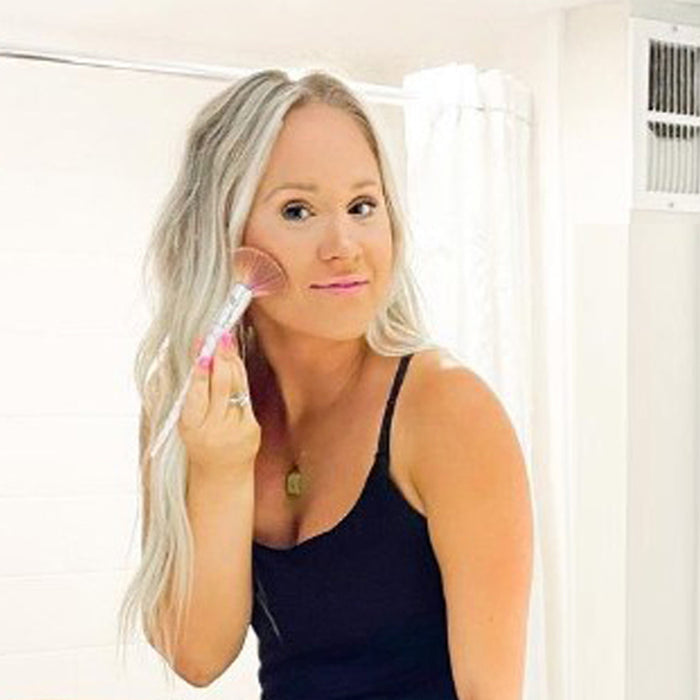 A woman in front of a mirror applying Demure Face & Body Bronzer