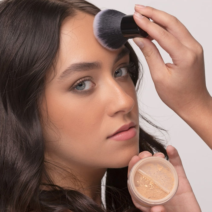 Woman applying Demure Medium Warm Foundation with a Kabuki brush and powder jar on a neutral background