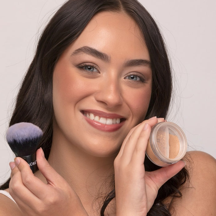 Woman holding a makeup brush and container of Demure Medium Warm Foundation powder against a neutral background