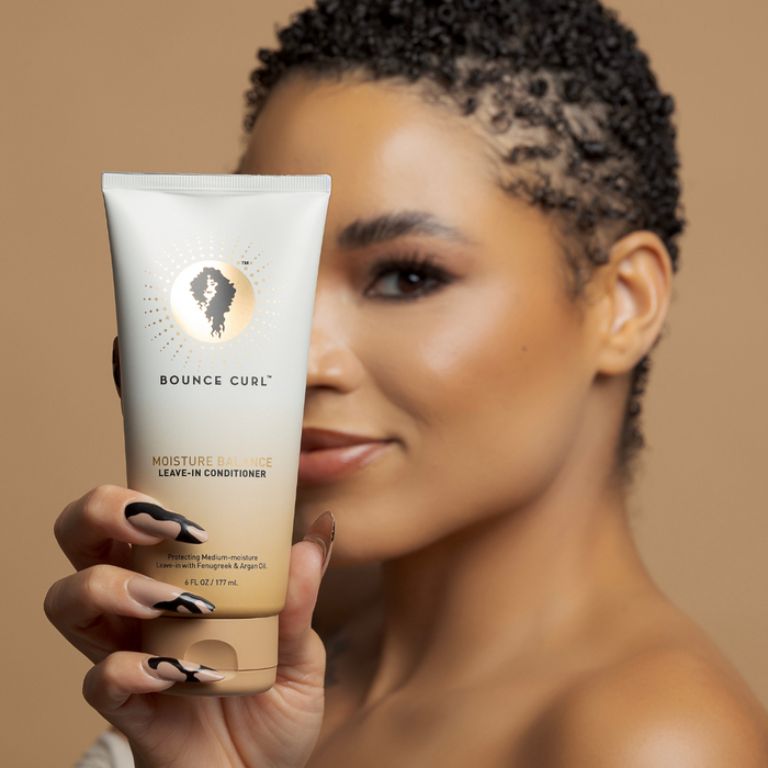 Person with short curls holding a Bounce Curl product tube against a warm tan background.