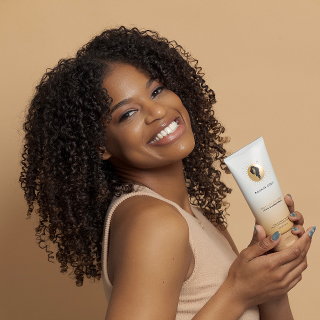 Woman with dark curly hair smiling and holding Bounce Curl’s Moisture Balance Leave-in Conditioner