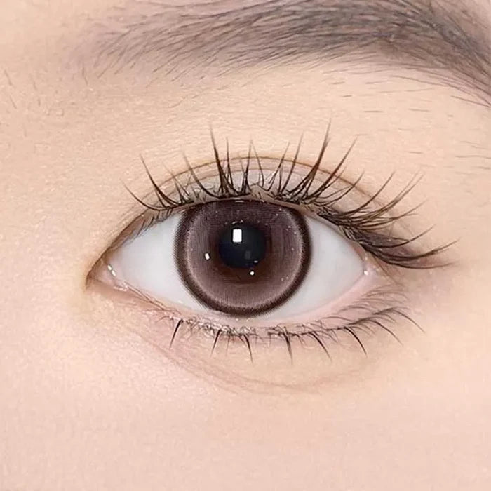 A close-up of a human eye with long lashes wearing Olola Meow Natural Angora Pink contacts by Olola, showing detailed iris texture and smooth, light skin around the eye.