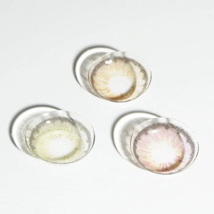 Three colored contact lenses—brown, green, and EyeCandys Pink Label Angel Grey—are arranged in a triangle on a white surface, each lens casting a faint shadow.