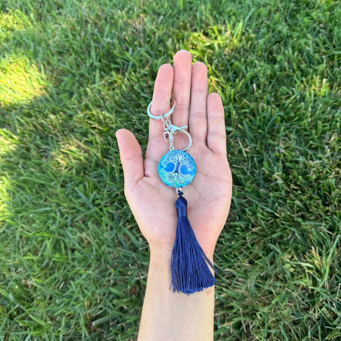 Hand holding a keychain with a blue tree of life design and tassel against a grass background