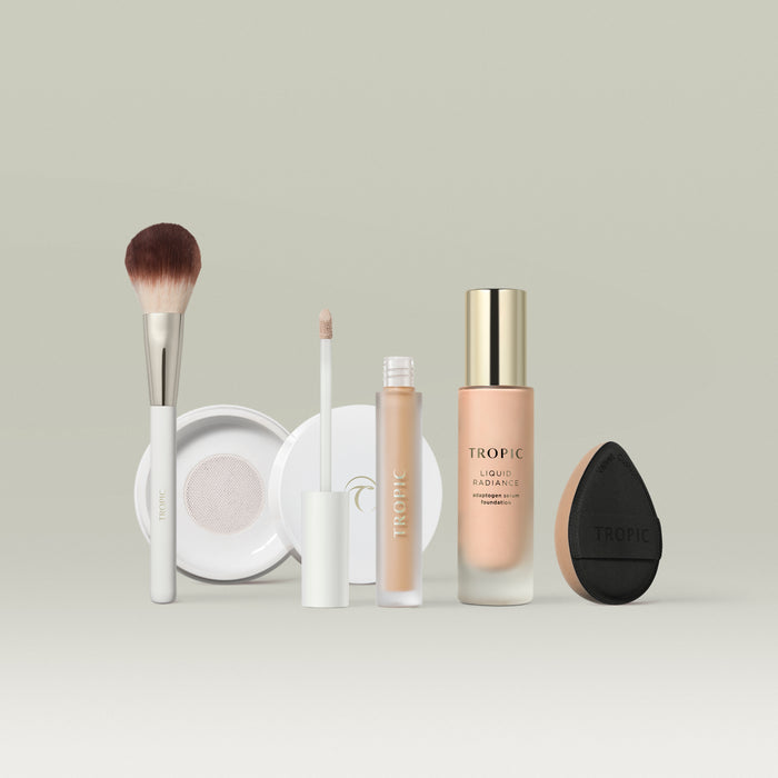 A set of Tropic makeup products in shade 3 arranged in a row against a soft neutral background. The lineup includes a fluffy makeup brush, a white compact, a liquid concealer with a doe-foot applicator, a bottle of Liquid Radiance foundation with a gold cap, and a black teardrop-shaped blending sponge.