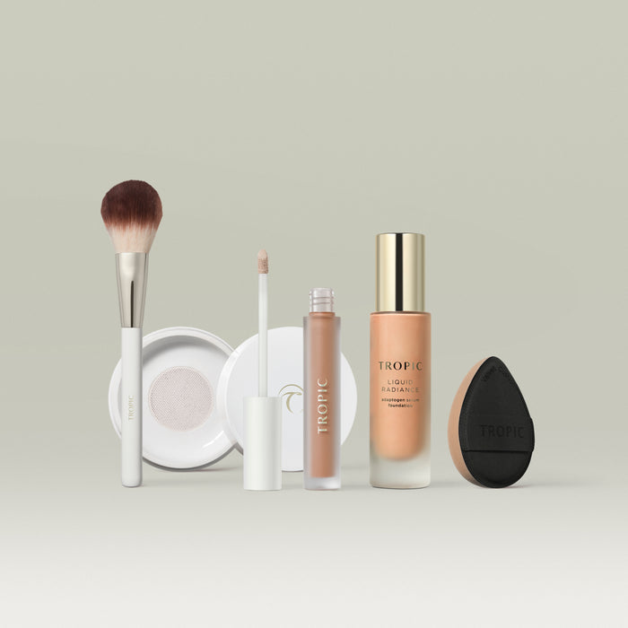 A set of Tropic makeup products in shade 6 arranged in a row against a soft neutral background. The lineup includes a fluffy makeup brush, a white compact, a liquid concealer with a doe-foot applicator, a bottle of Liquid Radiance foundation with a gold cap, and a black teardrop-shaped blending sponge.
