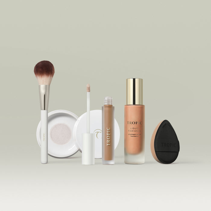 A set of Tropic makeup products in shade 7 arranged in a row against a soft neutral background. The lineup includes a fluffy makeup brush, a white compact, a liquid concealer with a doe-foot applicator, a bottle of Liquid Radiance foundation with a gold cap, and a black teardrop-shaped blending sponge.