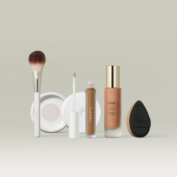 A set of Tropic makeup products in shade 8 arranged in a row against a soft neutral background. The lineup includes a fluffy makeup brush, a white compact, a liquid concealer with a doe-foot applicator, a bottle of Liquid Radiance foundation with a gold cap, and a black teardrop-shaped blending sponge.
