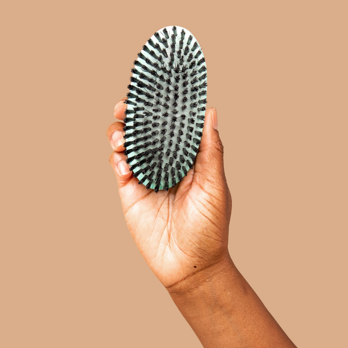Hand holding an oval hairbrush with black and mint bristles on a tan background.