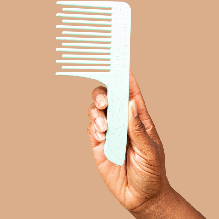 Hand holding a white wide‑tooth comb with handle on a tan background.