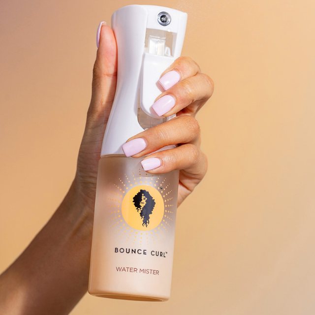 Hand holding Bounce Curl water mister spray bottle with white trigger, warm background.